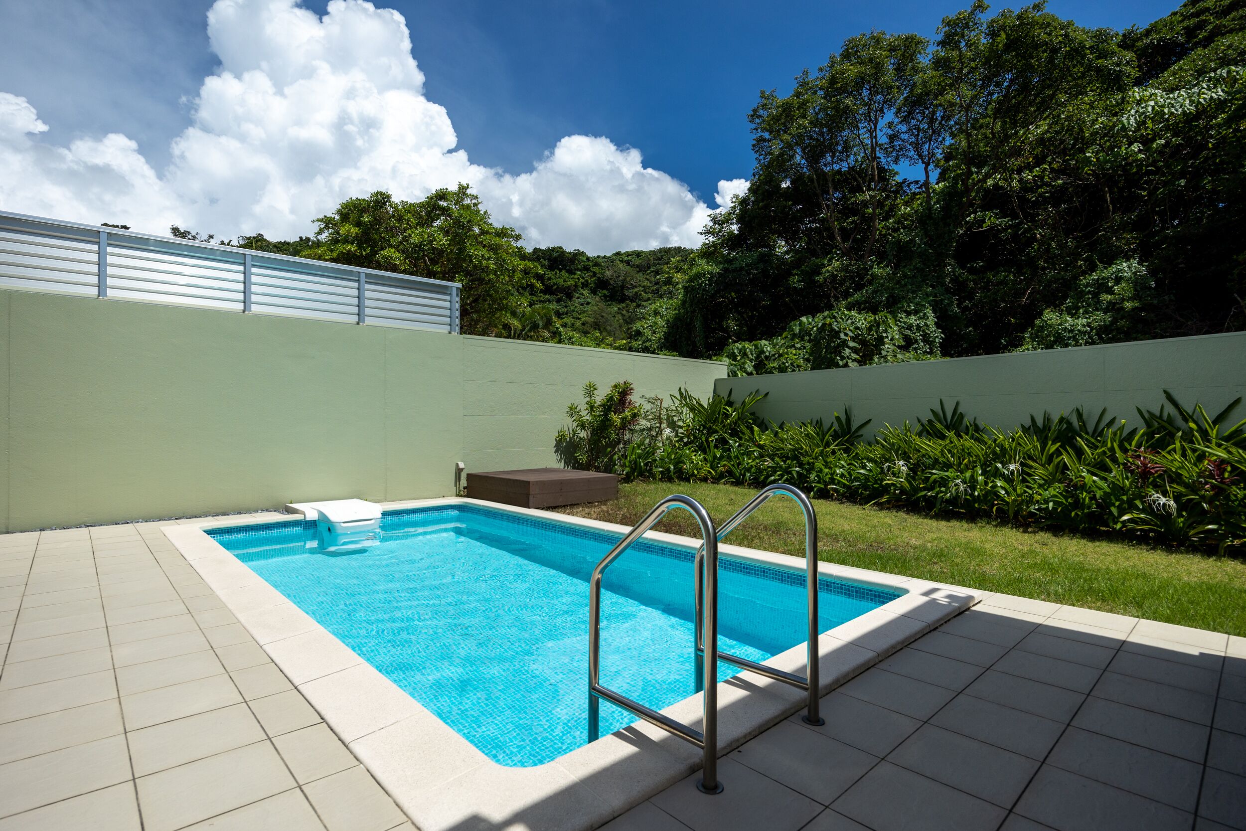 Piscina rectangular con una escalera metálica, rodeada por un suelo de baldosas y un muro bajo.