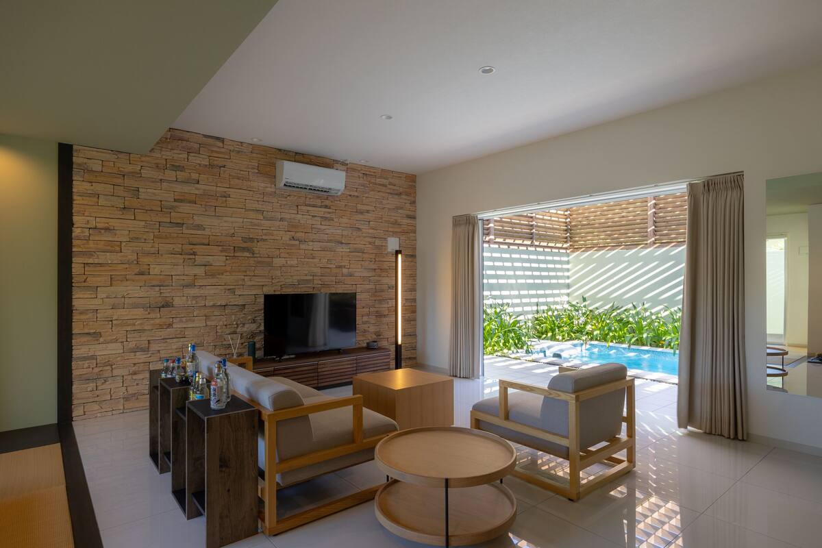 Una sala de estar moderna con una pared de piedra, un televisor de pantalla plana, muebles de madera y vista a una piscina.