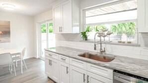 A modern kitchen with white cabinetry, a marble countertop, a stainless steel sink, and a window with a view of greenery. - Wow! Beachside Glam 3br wPool, Walk to Park, Beach (Satellite Beach)