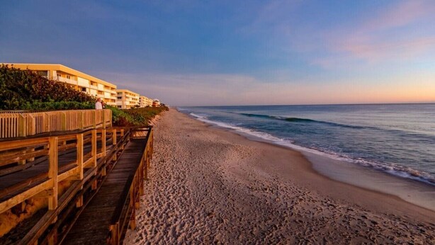 A beach with a wooden boardwalk leading to a building with multiple balconies. - Wow! Beachside Glam 3br wPool, Walk to Park, Beach (Satellite Beach)