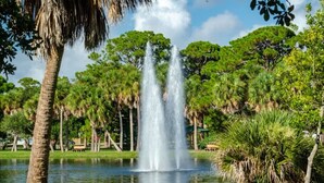 A park with a central fountain, surrounded by trees and a calm body of water. - Wow! Beachside Glam 3br wPool, Walk to Park, Beach (Satellite Beach)