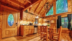 Une cuisine rustique aménagée dans une cabane en rondins, avec des meubles en bois, une table à manger et une grande fenêtre donnant sur une forêt.