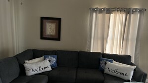 A living room with a dark grey sectional sofa, two 'welcome' pillows, a framed artwork on the wall, and sheer curtains. - Relax On The Coast (Foley)