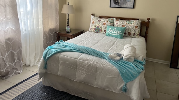A neatly made bed with a white comforter and a teal throw blanket, a bedside table with a lamp, and a framed abstract painting on the wall. - Relax On The Coast (Foley)