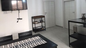 A modern, minimalist room with a flat-screen TV mounted on the wall, a dining table with chairs, and a small shelf with kitchen items. - TRAMONTANA apartment with whirlpool shower in the room (Taranto)