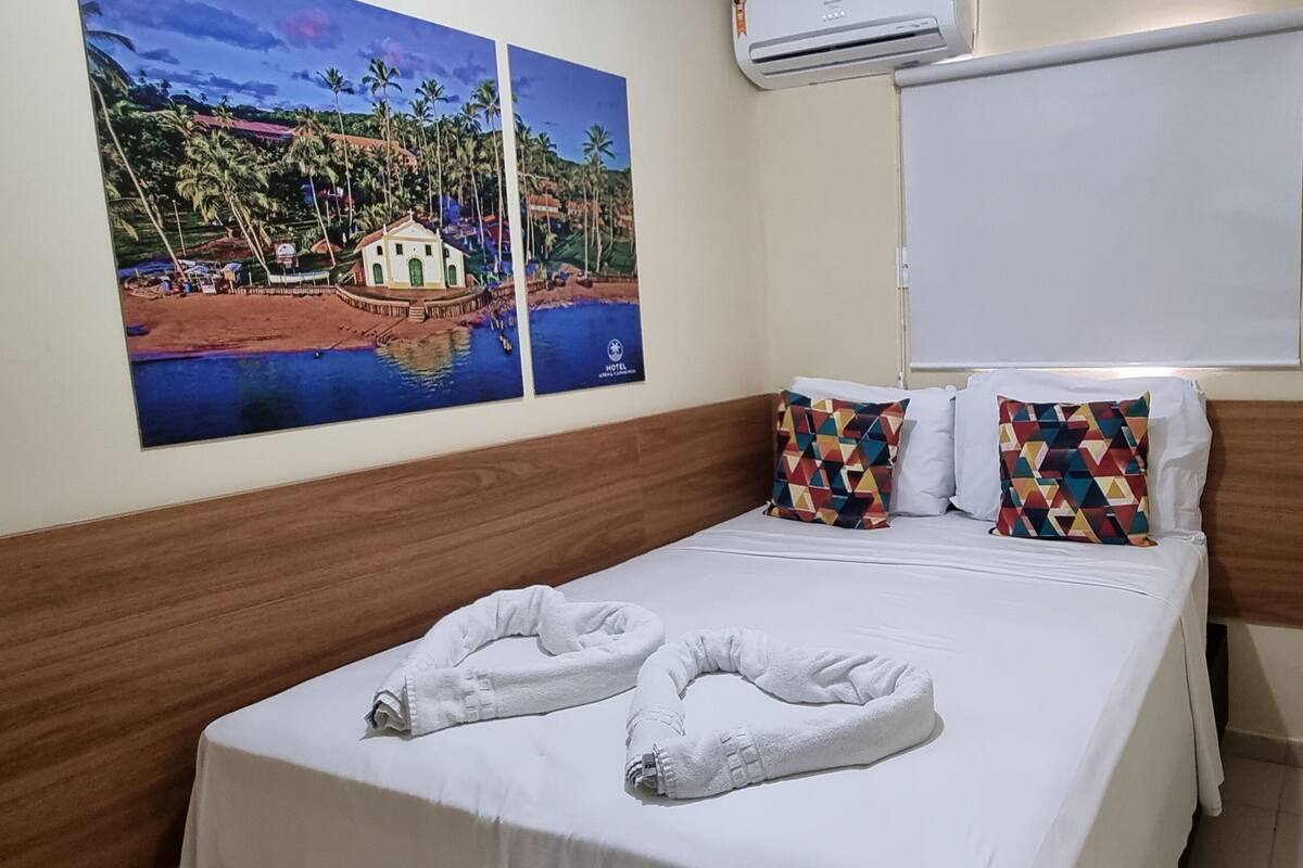 Une chambre d’hôtel avec un lit, deux oreillers, un tableau représentant un paysage côtier et une fenêtre avec un store.
