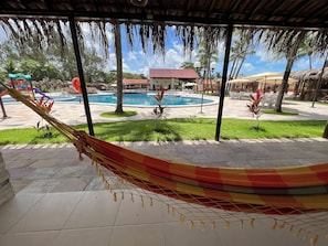 A hammock under a thatched roof, a swimming pool, and a building with a red roof in the background. - Hotel Arena Carneiros By AFT (Tamandaré)