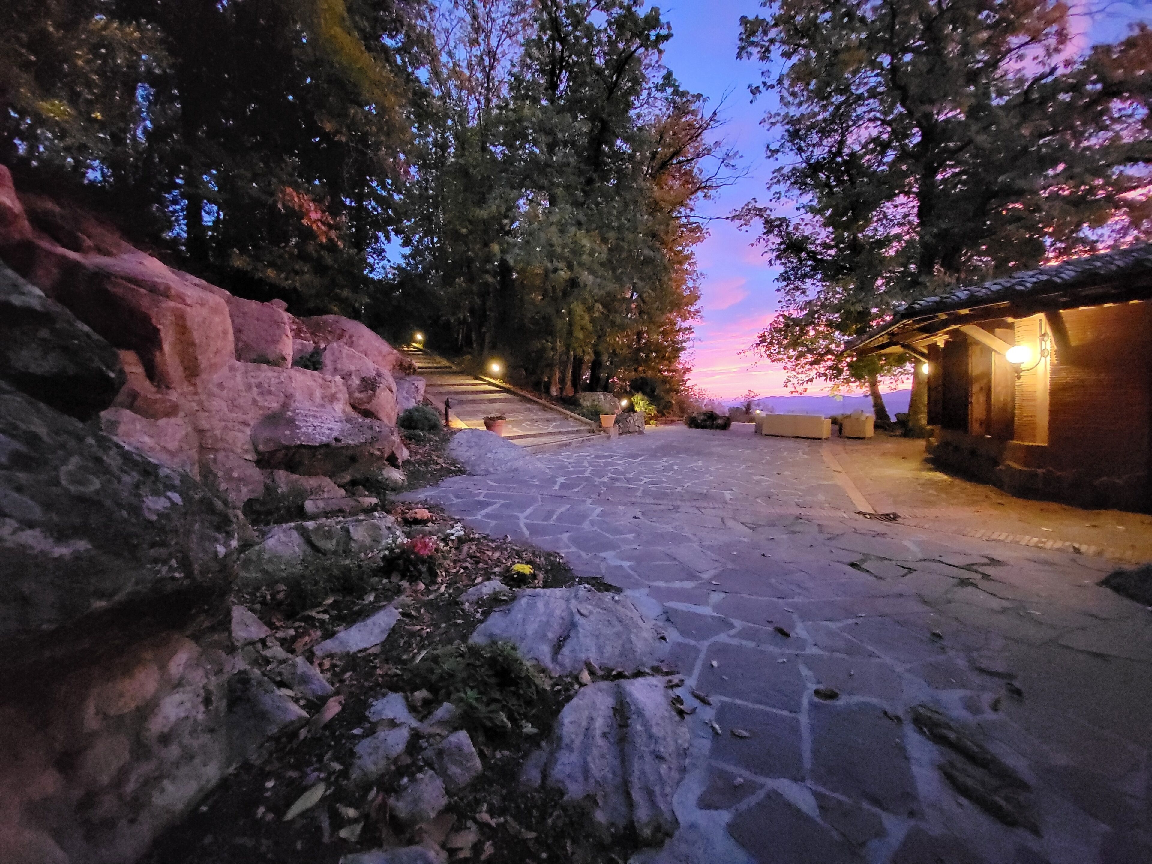 A stone pathway leading to a building with a tiled roof, surrounded by trees and a rocky landscape.