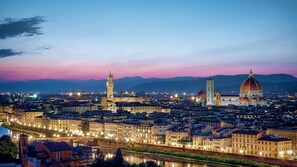 Une vue d’une ville au crépuscule, avec des bâtiments illuminés, une rivière et des éléments architecturaux historiques remarquables, notamment une tour et un dôme.