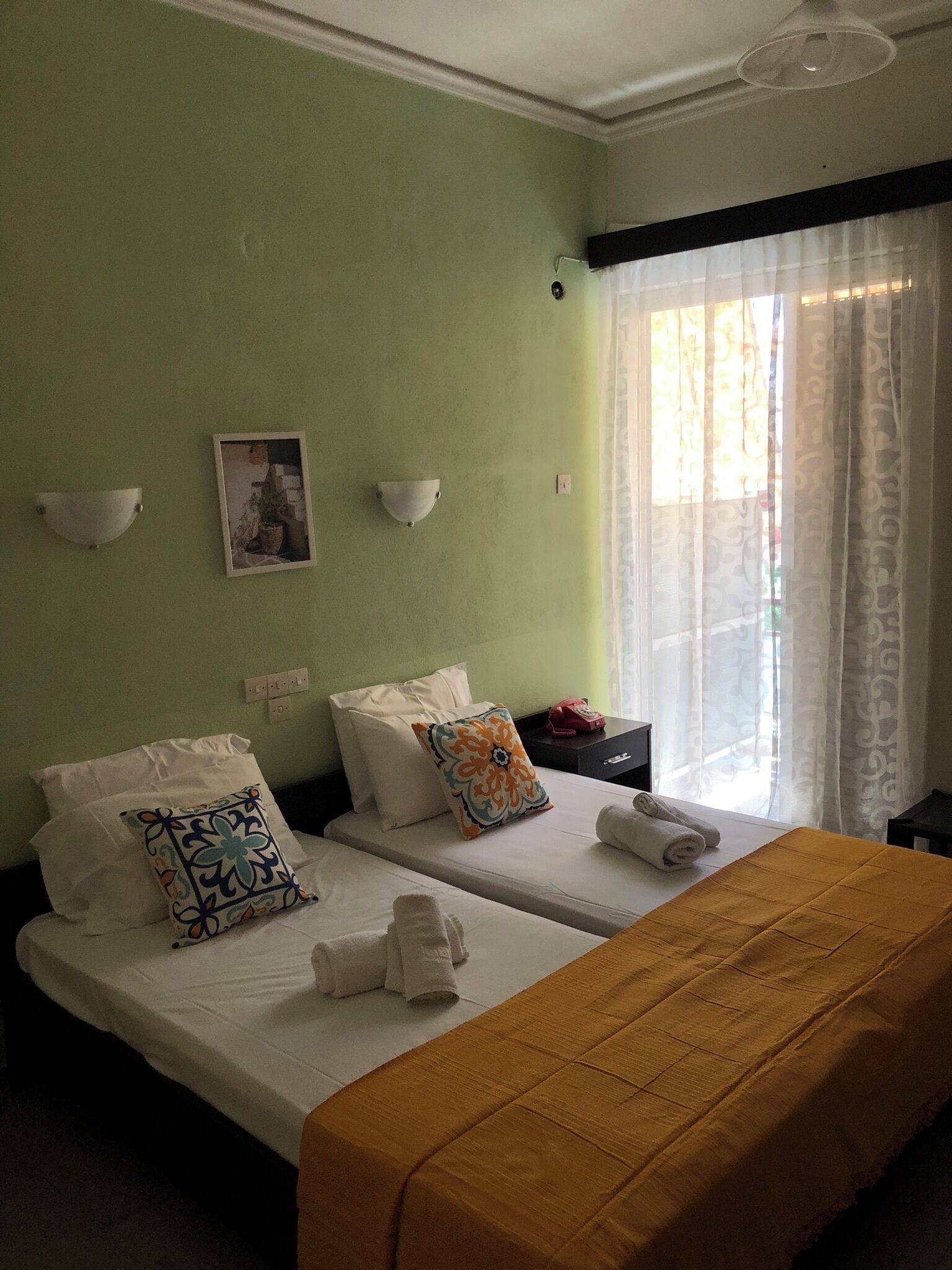 Habitación de hotel con dos camas, un ventanal con cortinas translúcidas, una lámpara de pared y una mesita de noche pequeña con teléfono.