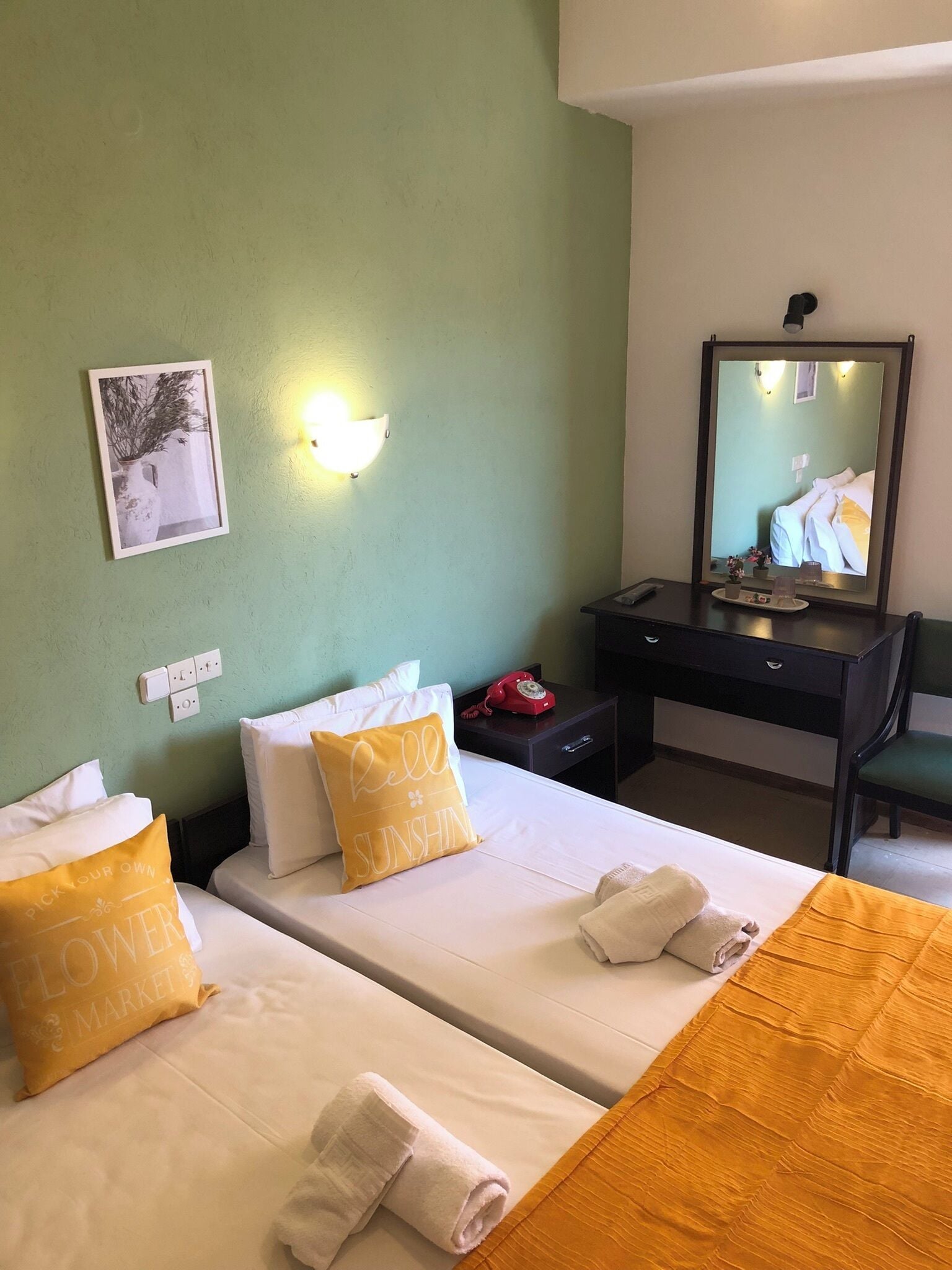 A hotel room with two beds, a desk, a mirror, and a wall-mounted light.