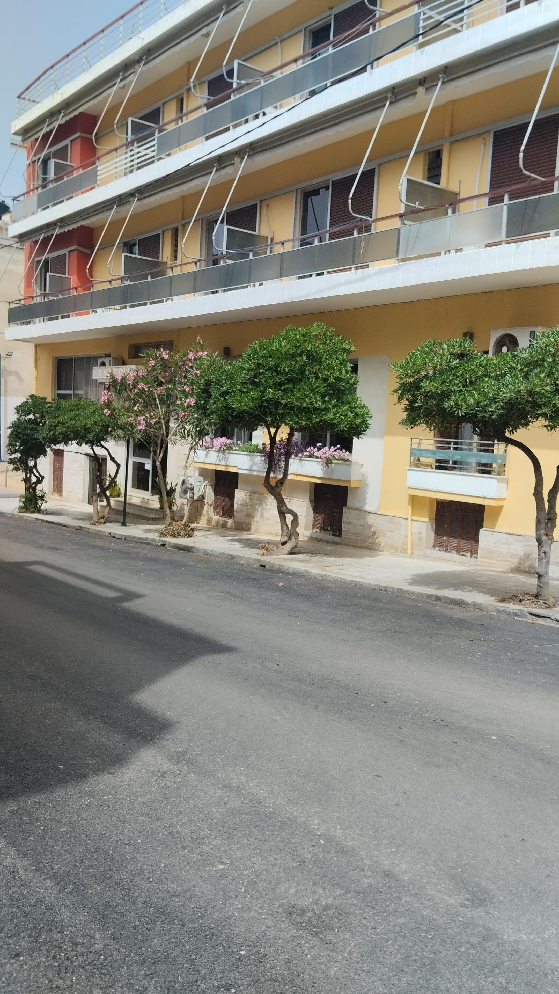 A multi-story building with balconies, a paved street, and small trees along the sidewalk.