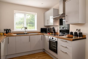 A modern kitchen with white cabinets, a built-in oven, a microwave, and a window with blinds. - Apartment 3 (Holyhead)
