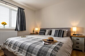 Ein ordentlich gemachtes Bett mit einer Karo-Decke, einem Strohhut, Sonnenbrille und einer Zeitschrift auf den Kissen. Zwei Nachttischlampen, ein Fenster mit Vorhängen und eine Heizung sind ebenfalls zu sehen.