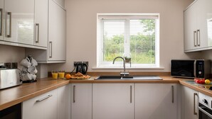 A modern kitchen with white cabinets, a wooden countertop, a sink, a microwave, and a window with blinds. - Apartment 3 (Holyhead)