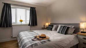 A neatly made bed with a striped blanket, a straw hat, and sunglasses on it. There is a window with curtains, a bedside table with a lamp, and a nightstand with a bottle and glasses. - Apartment 3 (Holyhead)