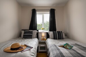 Ein Schlafzimmer mit zwei Betten, einem Nachttisch mit Lampe, einem Fenster mit Vorhängen und einem Hut auf dem Bett links.