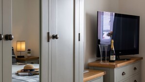 A hotel room with a bed, a bedside table with a lamp, a television, and a minibar with a bottle of champagne and glasses. - Apartment 3 (Holyhead)