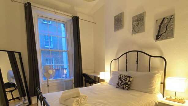 A neatly made bed with white linens and a rolled towel, a bedside table with a lamp, a black metal headboard, a window with curtains, and a radiator.