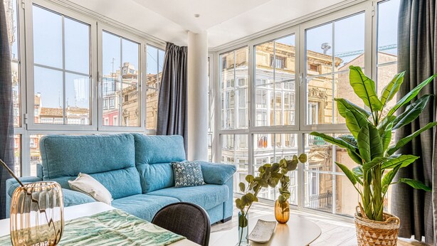 A modern living room with a blue sofa, a dining table, and large windows offering city views. - Limehome Haro Calle de la Vega (Haro)