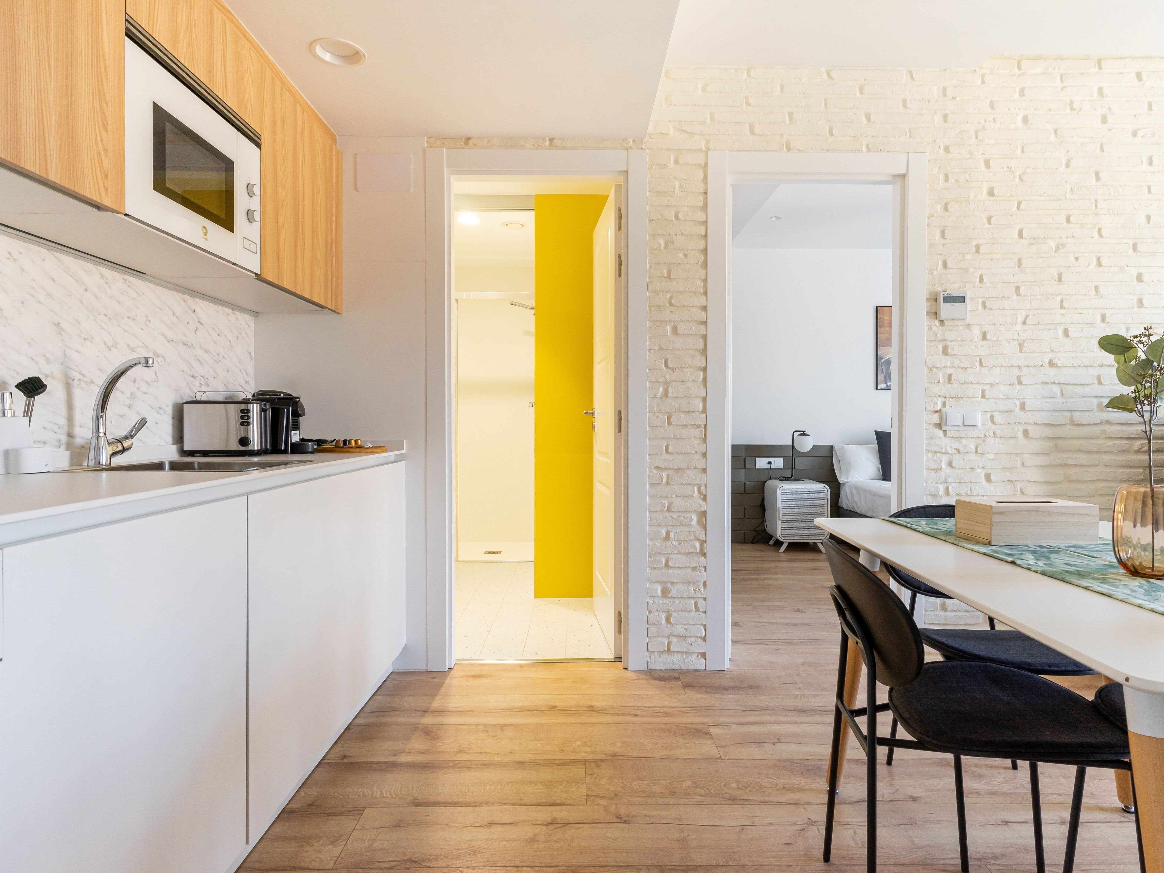 A modern kitchen with white cabinets, a microwave, and a toaster. There is a wooden dining table with chairs and a yellow accent wall.