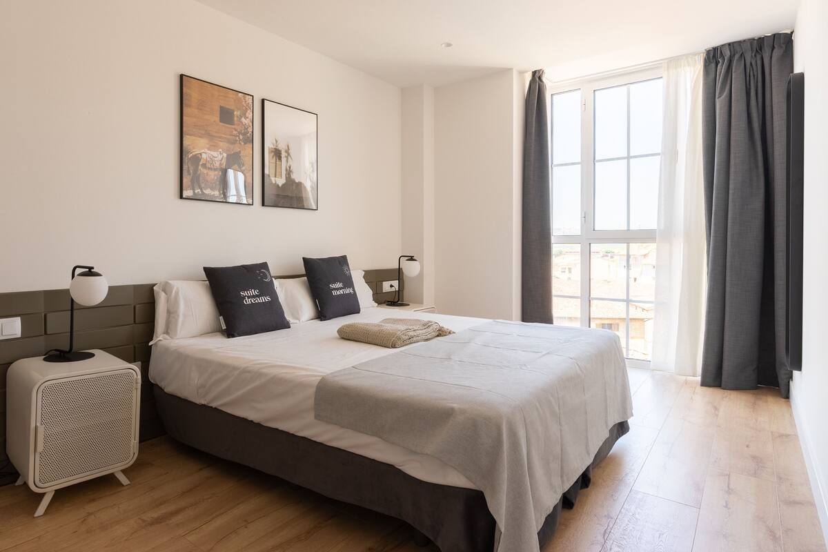 A bedroom with a bed, two pillows, a bedside table, a lamp, a radiator, and a window with curtains.