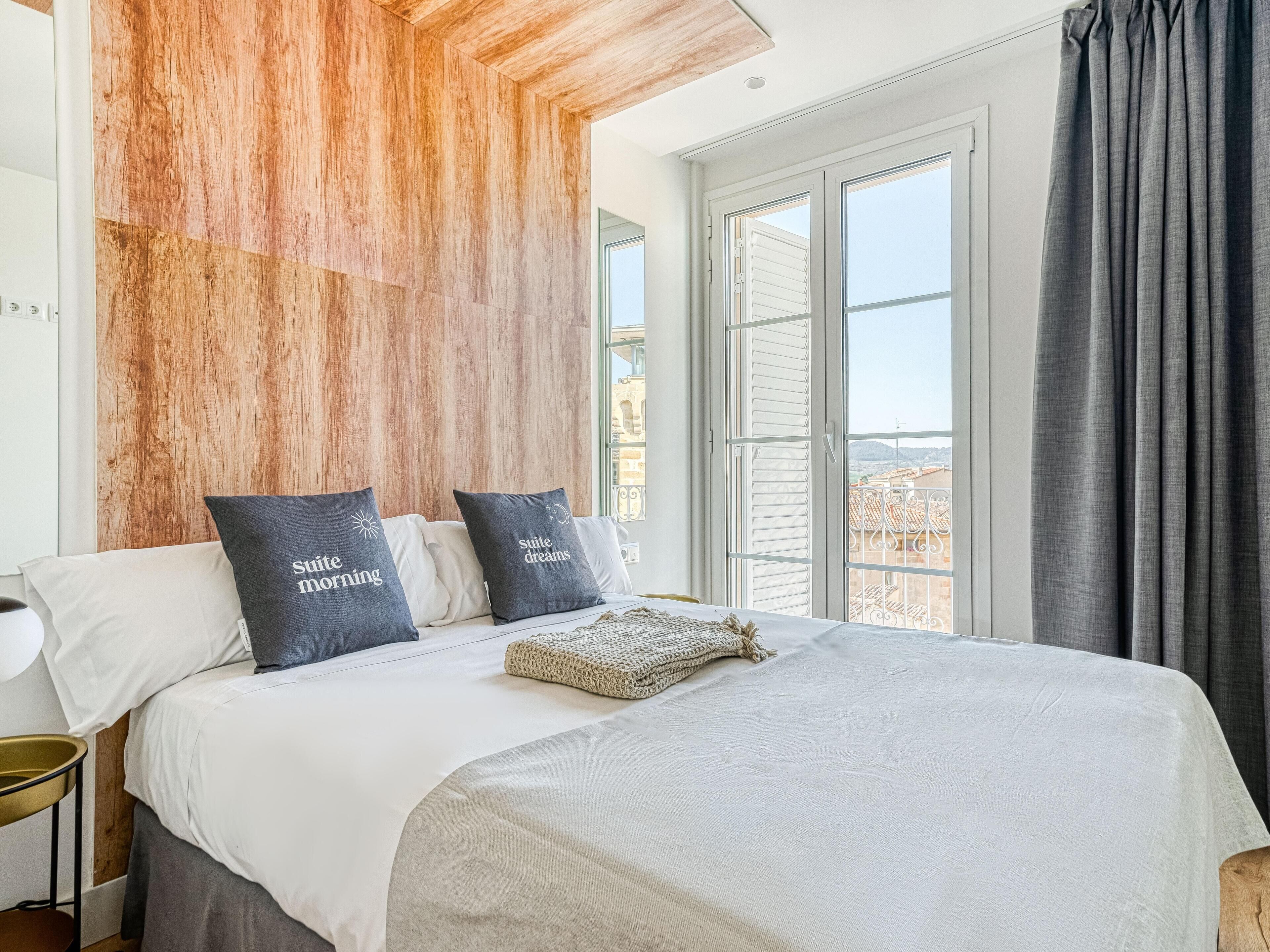 Un dormitorio con una cama grande, dos almohadas con texto, una manta tejida y vistas a edificios a través de la ventana.