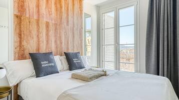 Un dormitorio con una cama grande, dos almohadas con texto, una manta tejida y vistas a edificios a través de la ventana.