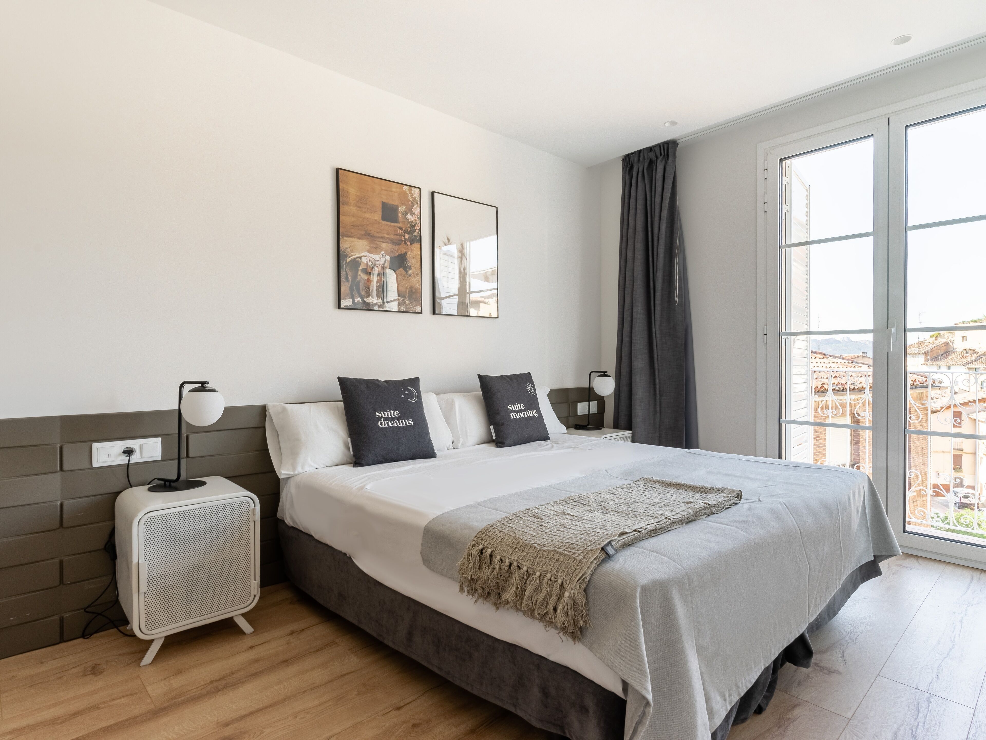 Habitación de hotel moderna con una cama grande, una mesita de noche con una lámpara, un altavoz pequeño y una ventana con vistas a edificios.