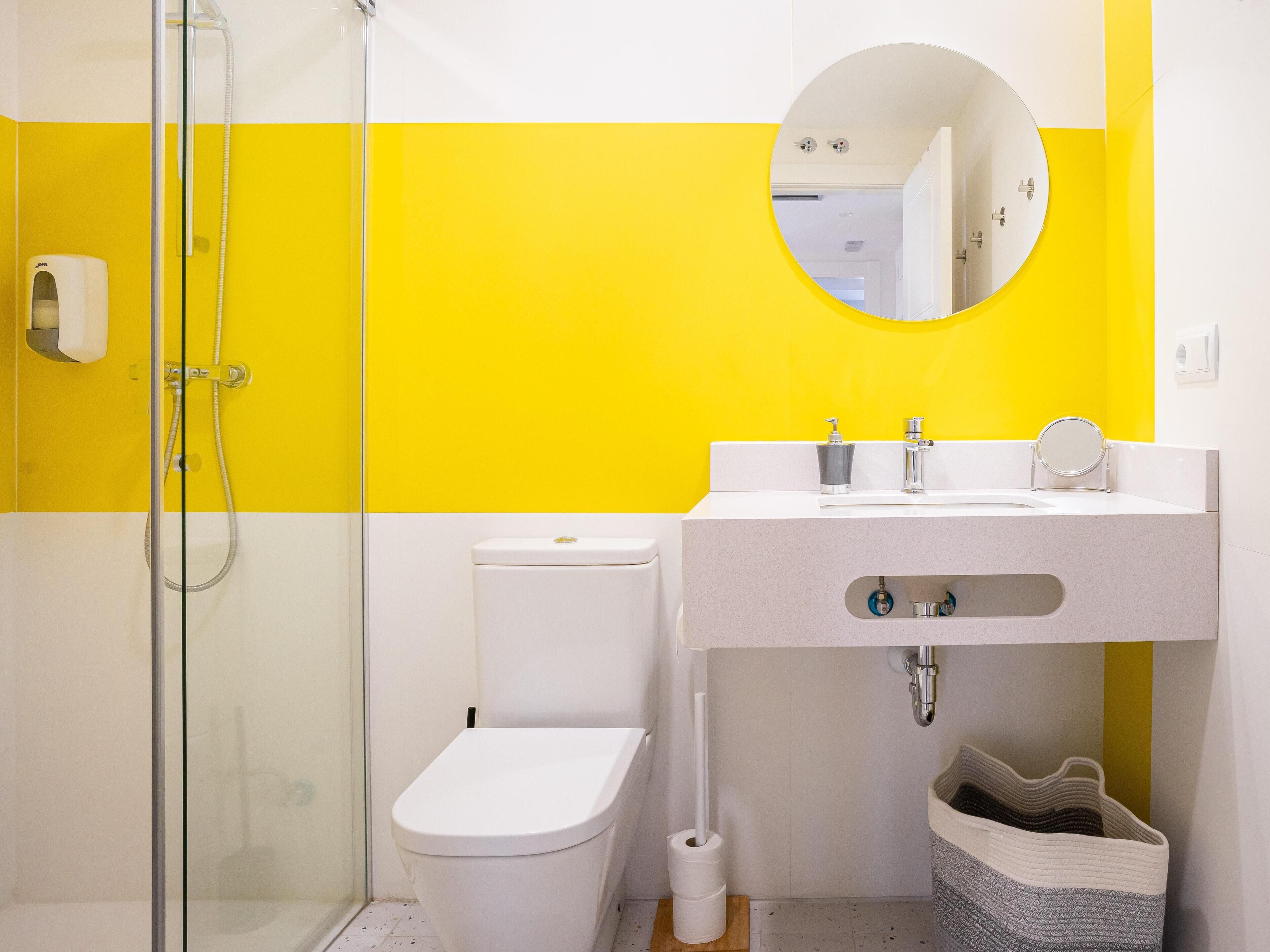 A bathroom with a glass shower enclosure, a toilet, a sink with a mirror, and a trash bin.