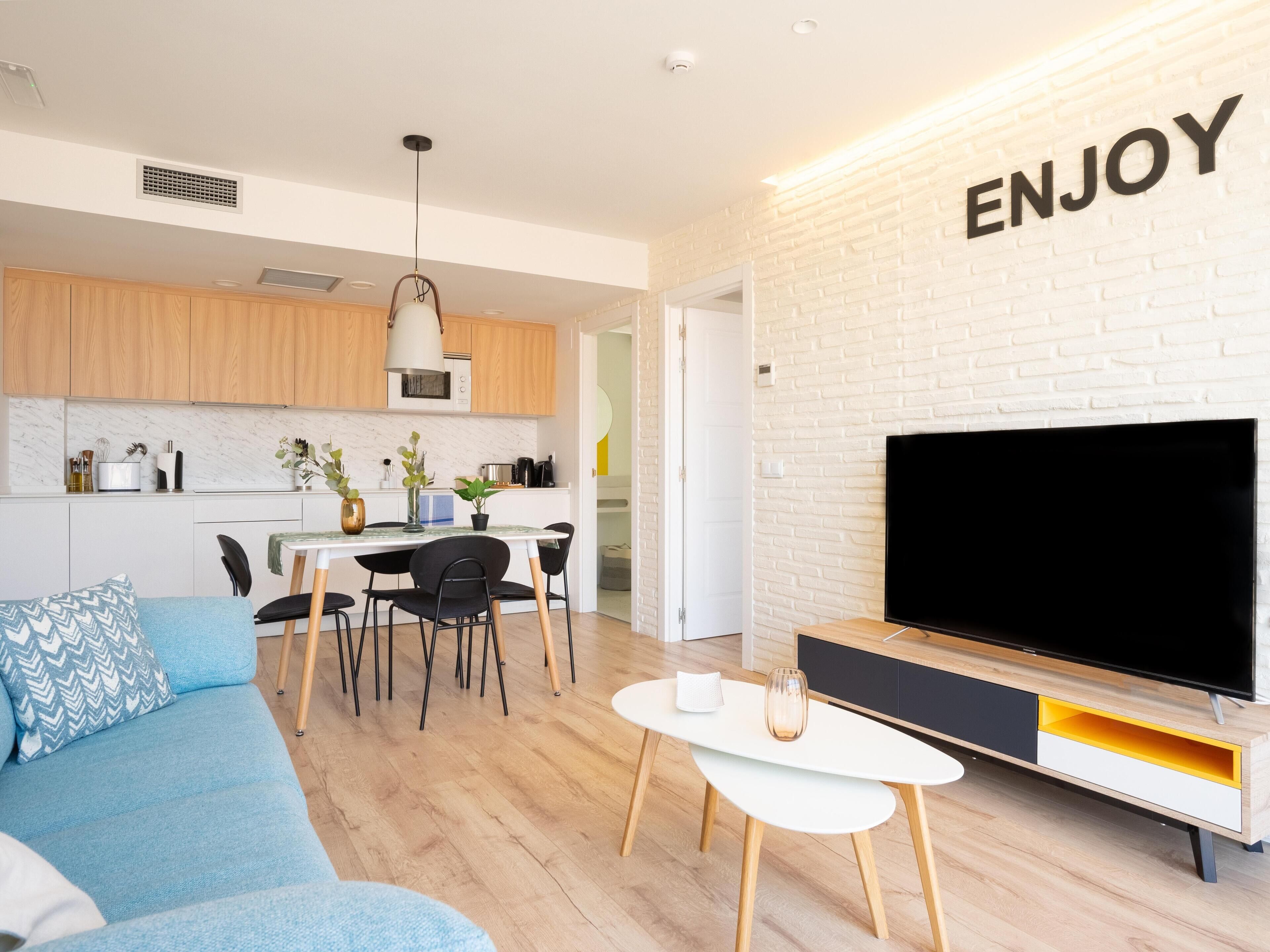 Una sala moderna con televisor de pantalla plana, un sofá azul, una zona de comedor con mesa y sillas, y una cocina al fondo.