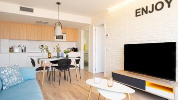 Una sala moderna con televisor de pantalla plana, un sofá azul, una zona de comedor con mesa y sillas, y una cocina al fondo.