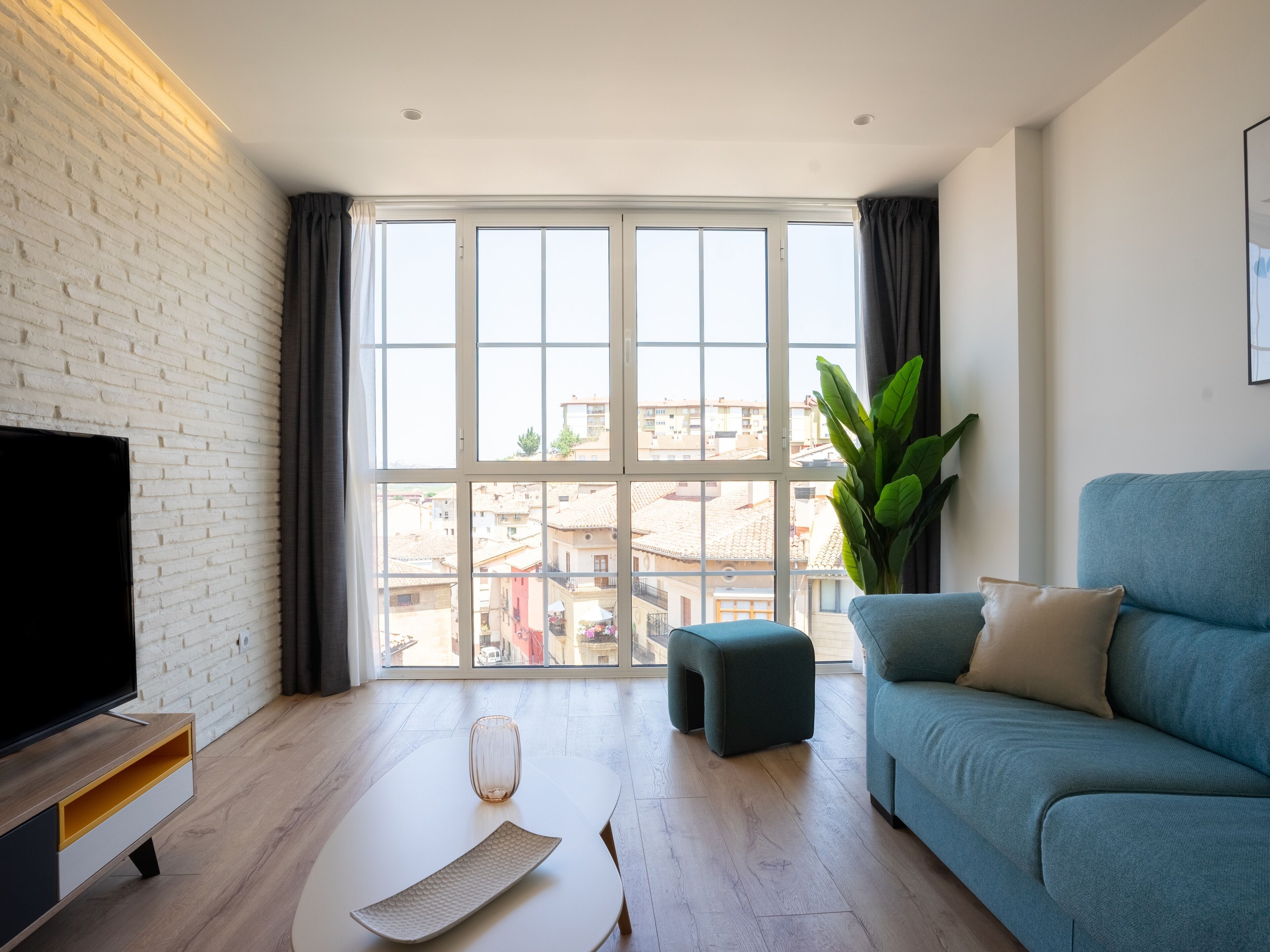 A modern living room with a large window, a flat-screen TV, a blue sofa, and a wooden coffee table.