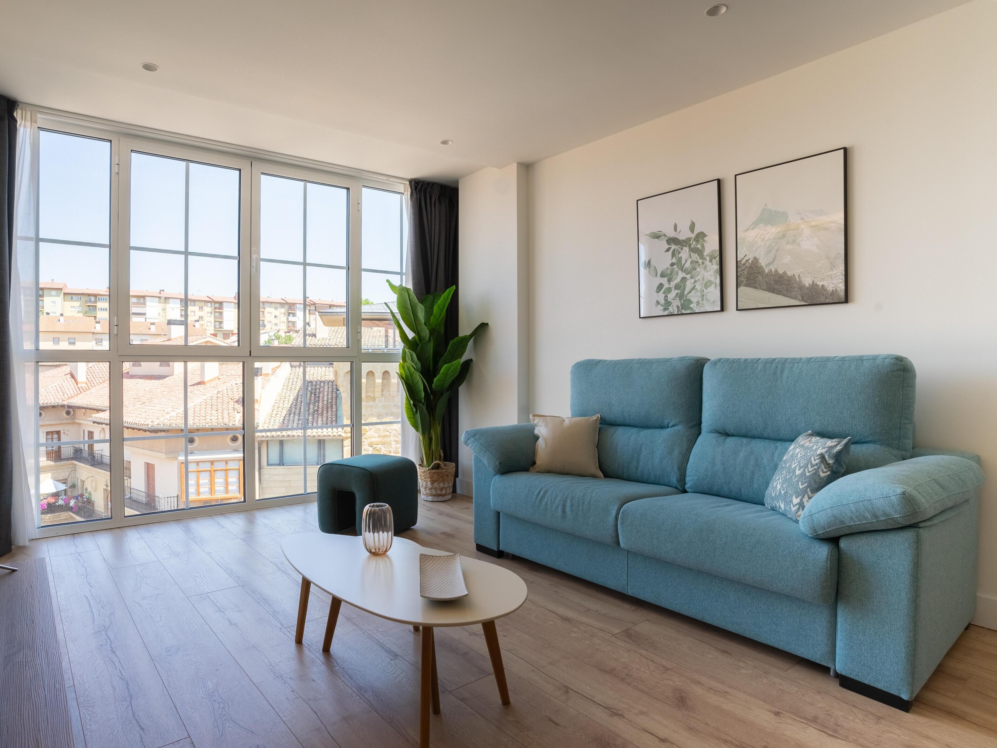 Una sala de estar moderna con un sofá azul, una mesa de centro, una planta en maceta y grandes ventanales con vistas a la ciudad.