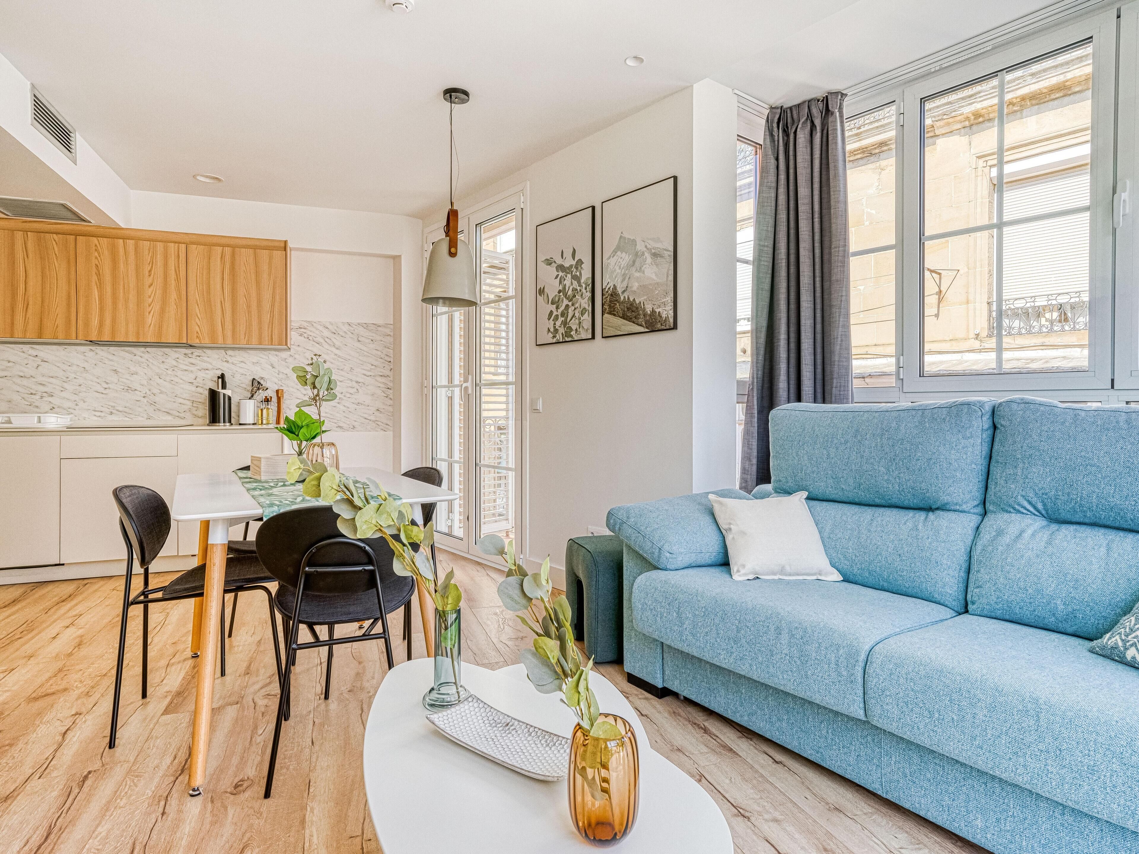 Una sala de estar moderna con un sofá azul, una mesa de comedor con sillas y una zona de cocina con armarios de madera.