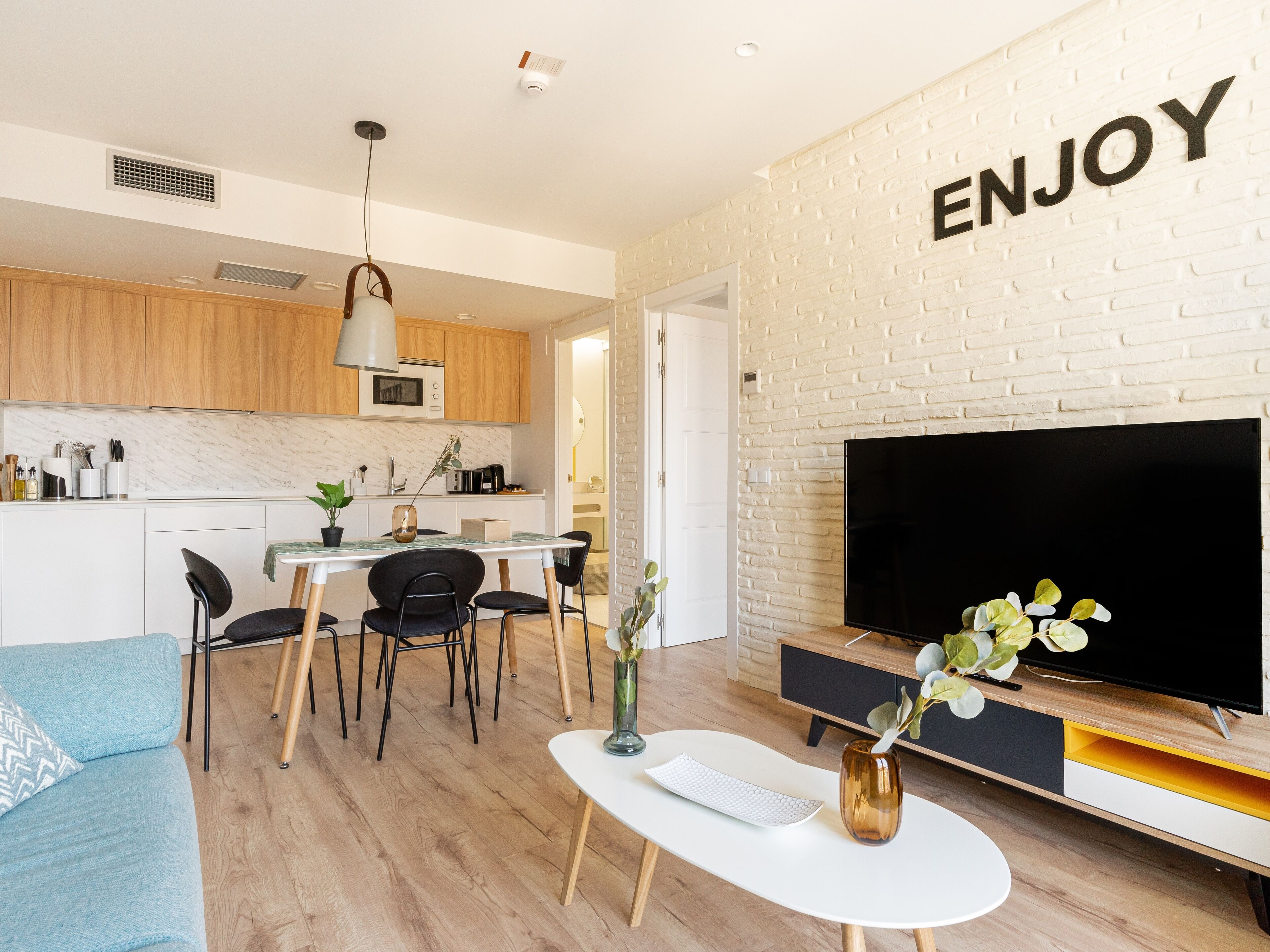 A modern living room with a flat-screen TV, a coffee table, and a sofa. There's a kitchen area with wooden cabinets and a white brick wall.