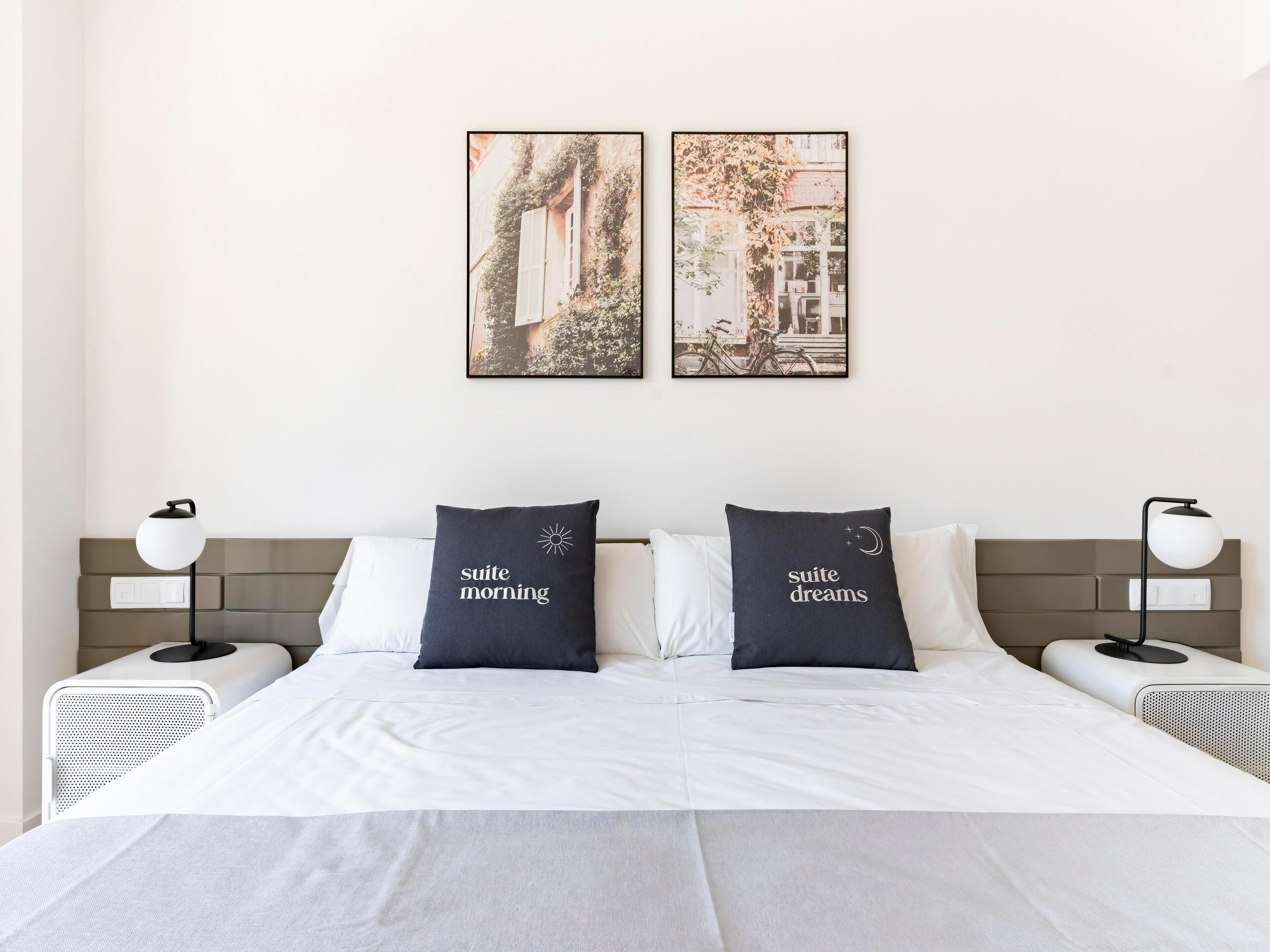 A hotel room with a bed, two pillows with text, a wall-mounted picture, and a nightstand with a lamp.