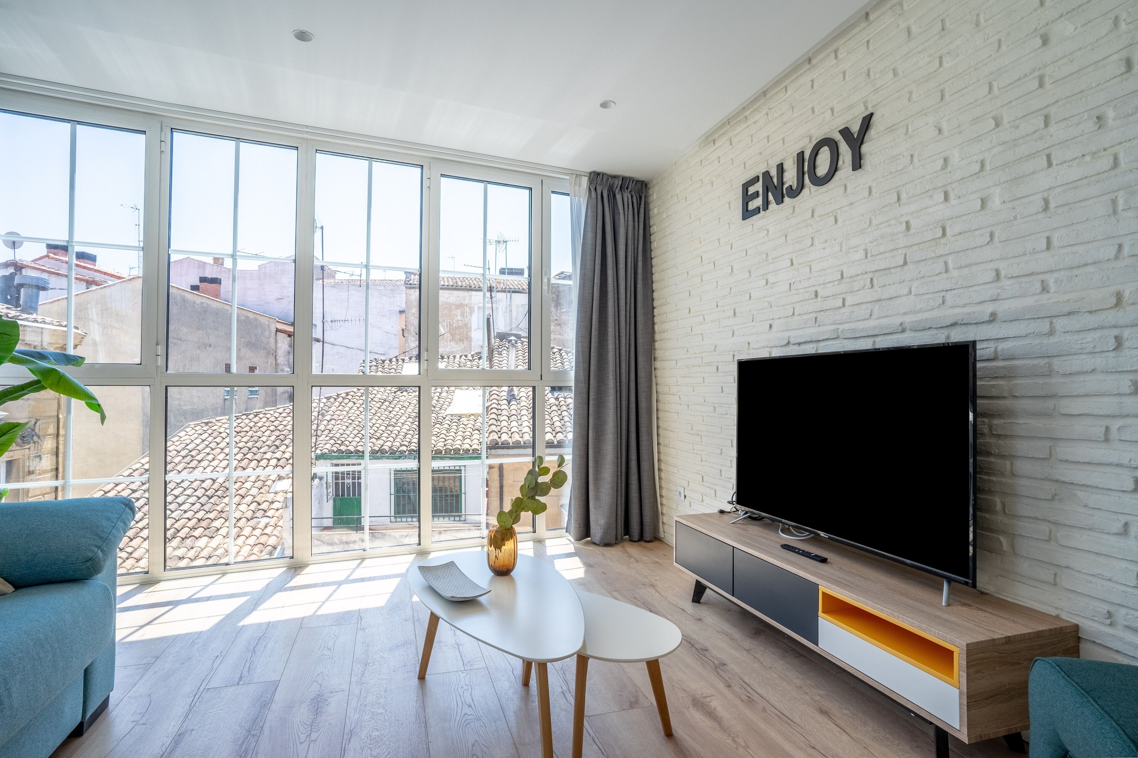 A modern living room with a large window, a flat-screen TV, and a 'ENJOY' sign on the wall.