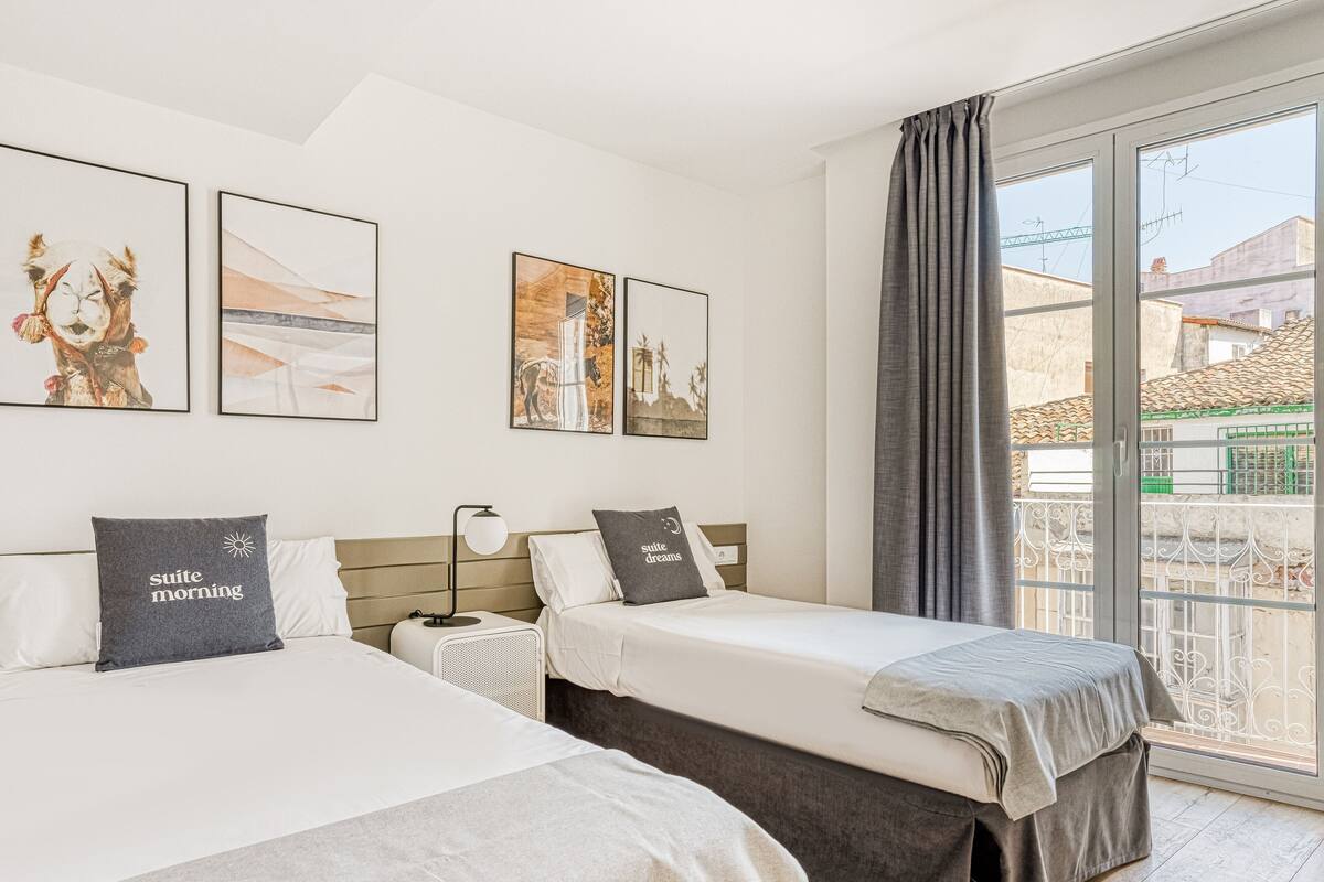 A hotel room with two beds, a large window with curtains, and three framed pictures on the wall.