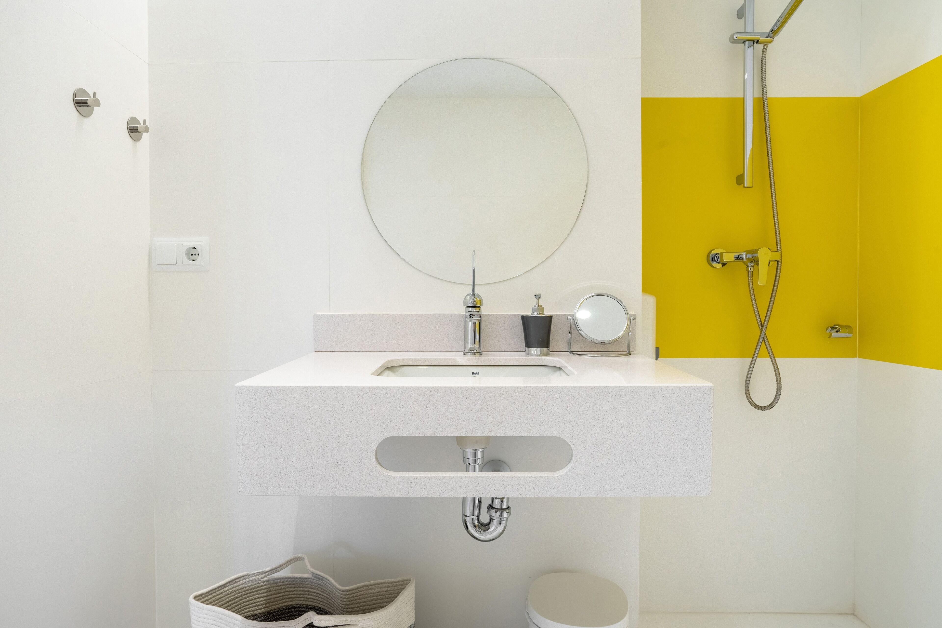 Un bany modern amb un lavabo blanc, un mirall rodó, un braç de dutxa portàtil i una paret d'accent groga.