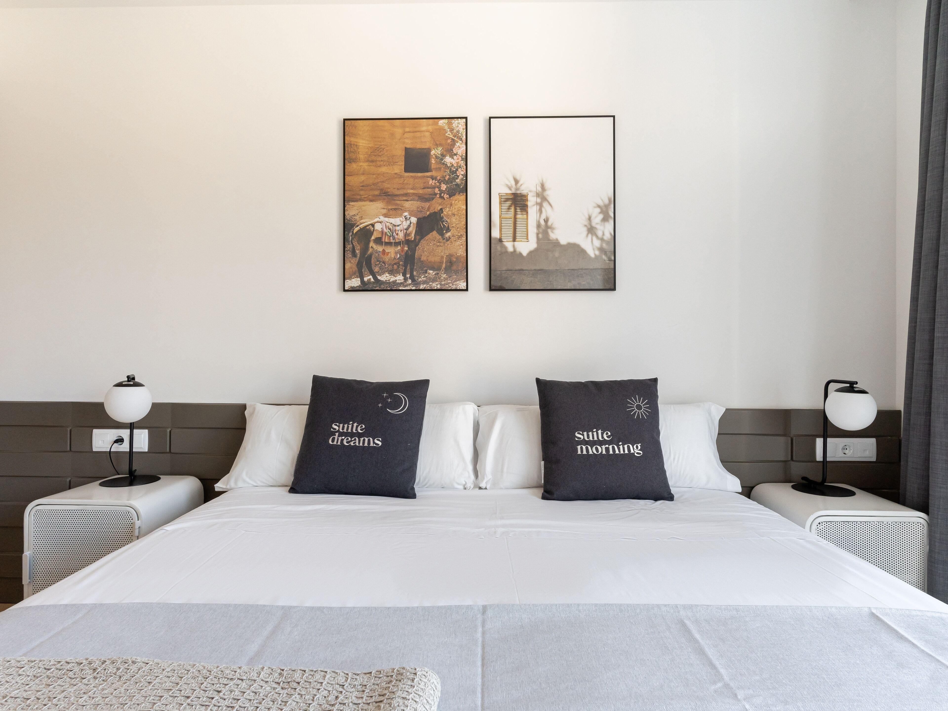 A hotel room with a large bed, two pillows with text, a nightstand with a lamp, and two wall-mounted artworks.