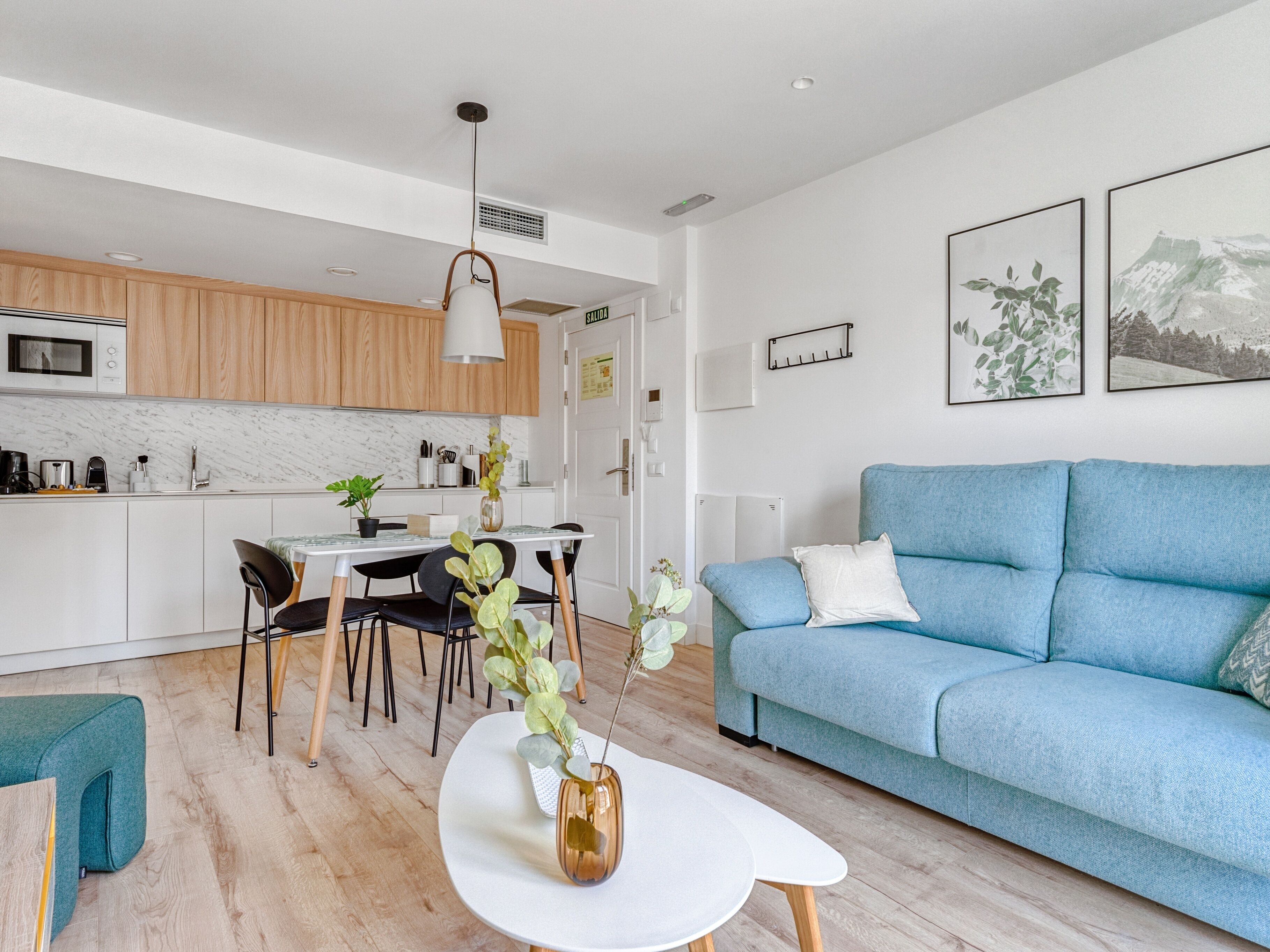 A modern living room with a blue sofa, a dining table with chairs, and a kitchen area with wooden cabinets and a white countertop.