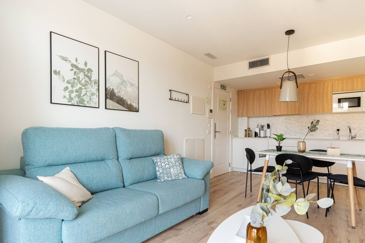 A modern living room with a blue sofa, a dining area with a white table, and a kitchen with wooden cabinets.