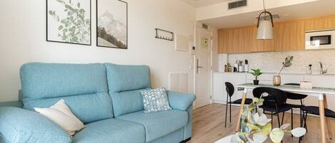 A modern living room with a blue sofa, a dining area with a white table, and a kitchen with wooden cabinets.