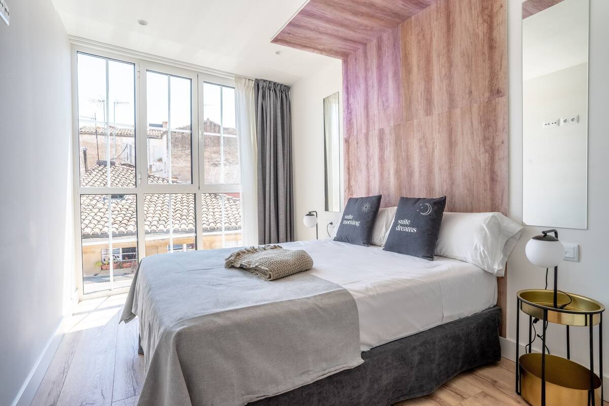 A modern bedroom with a large bed, a view of the city through a window, and a unique wooden accent wall.