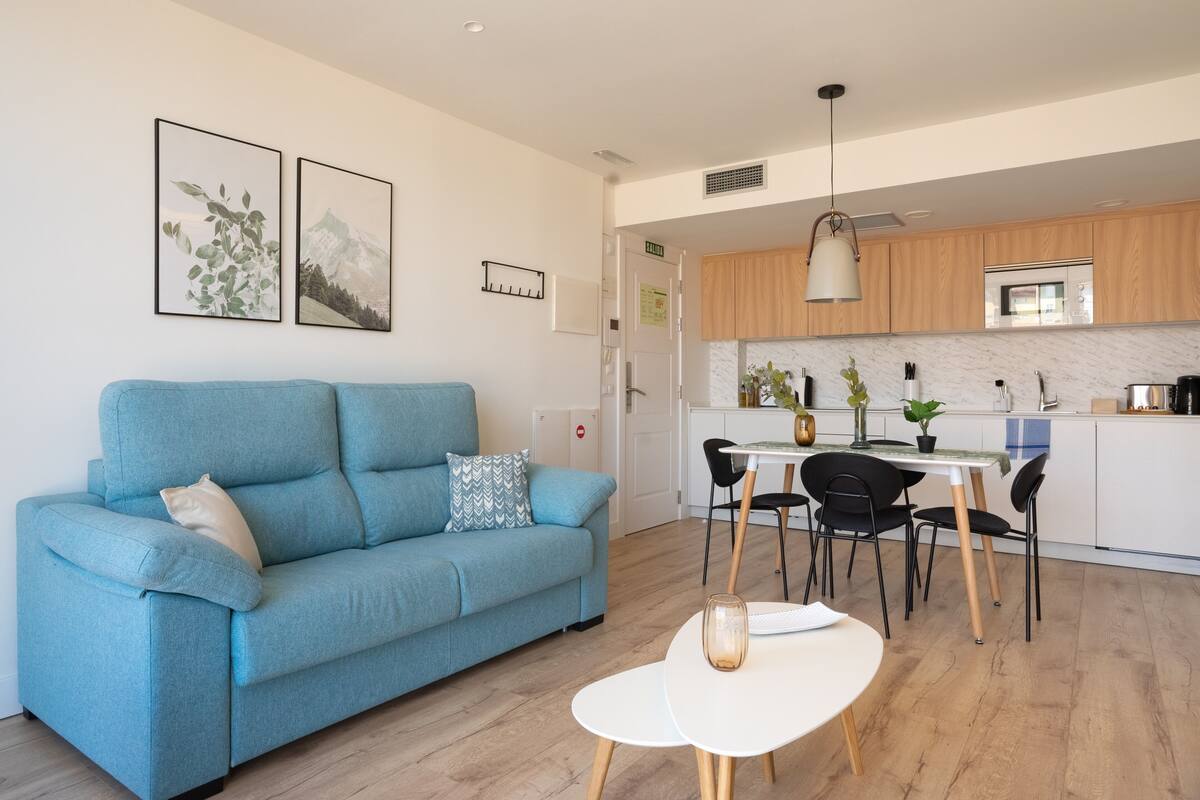 A modern living room with a blue sofa, a small white table, and a kitchen area with wooden cabinets and a white countertop.