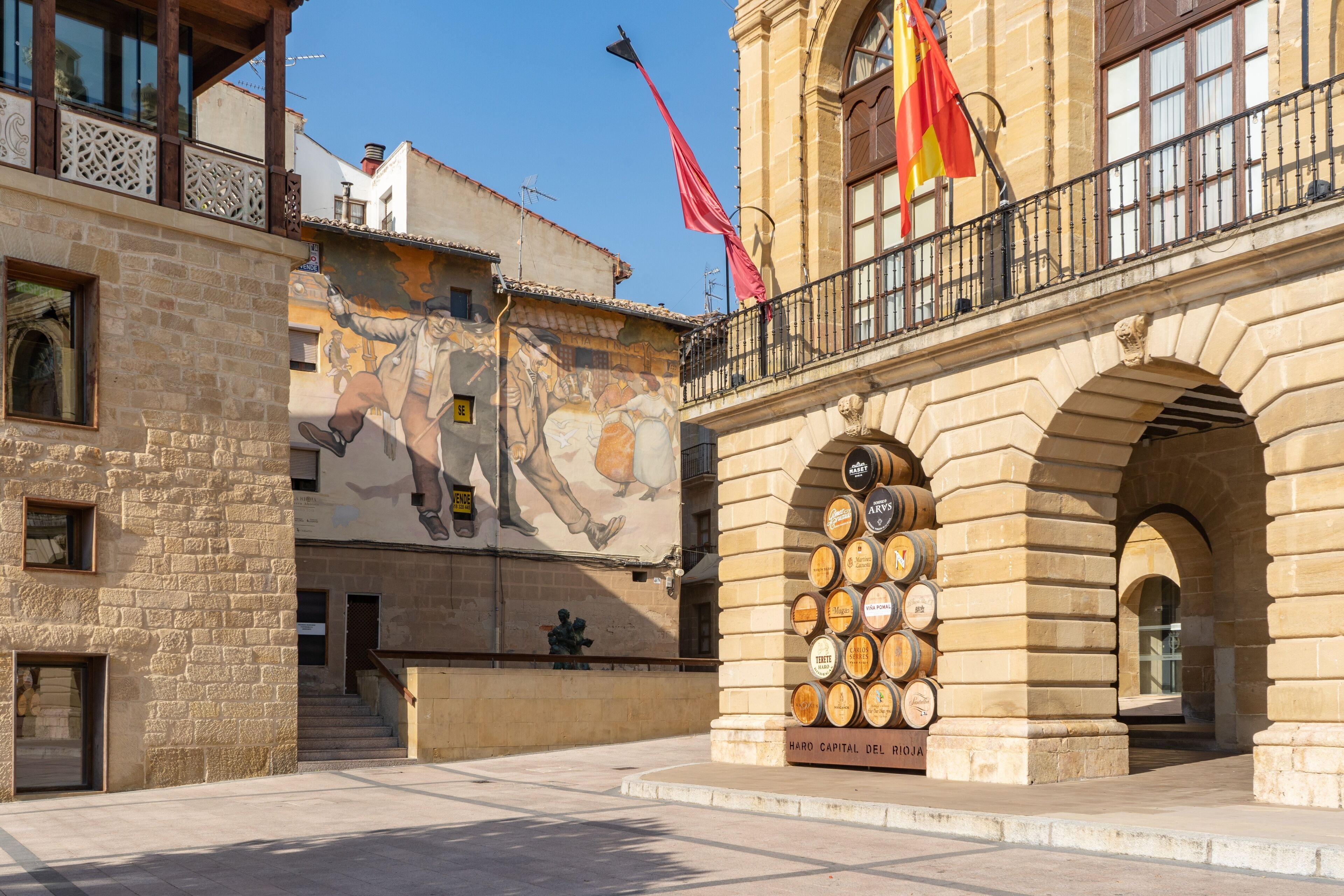 A historic building with a mural, a balcony with flags, and a display of wine barrels.