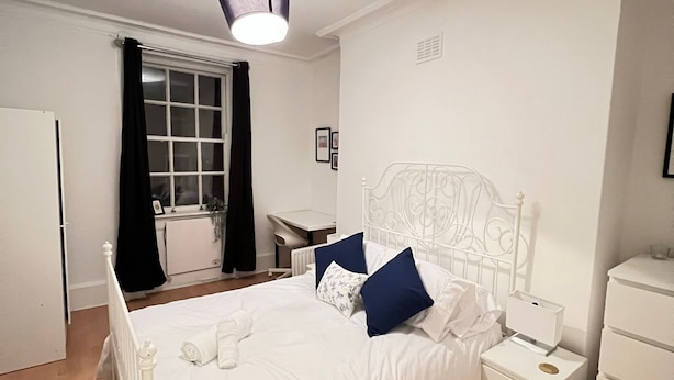 A bedroom with a white bed, a nightstand, a dresser, and a window with curtains. - Cosy 2-bed flat in the heart of SoHo! (London)