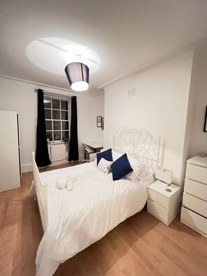 Quarto com cama branca, mesa de cabeceira, cômoda e janela com cortinas.
