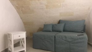 Una habitación pequeña y acogedora con una pared de piedra, un sofá gris con cojines y una mesita de noche blanca con cajón.